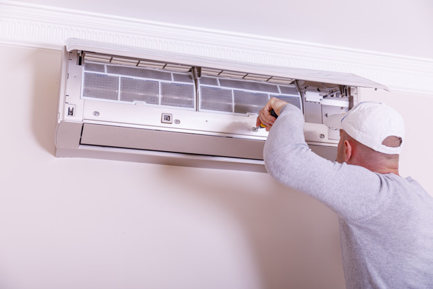 A technician is working on a wall-mounted air conditioning unit, using a screwdriver to access its internal components. The air conditioner is installed in a light-coloured room, highlighting the technician's focus on maintenance or repair.